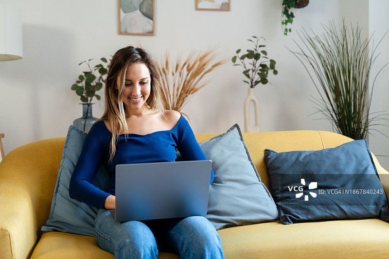 笑容灿烂的年轻女子在家中坐在沙发上使用笔记本电脑图片素材