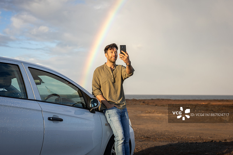 一名中年男游客在沙滩上自拍，身后是天空中出现的一道彩虹图片素材