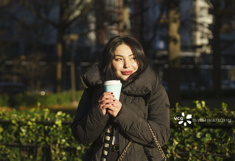 年轻女子身穿黑色外套，拿着一杯咖啡，在户外微笑图片素材