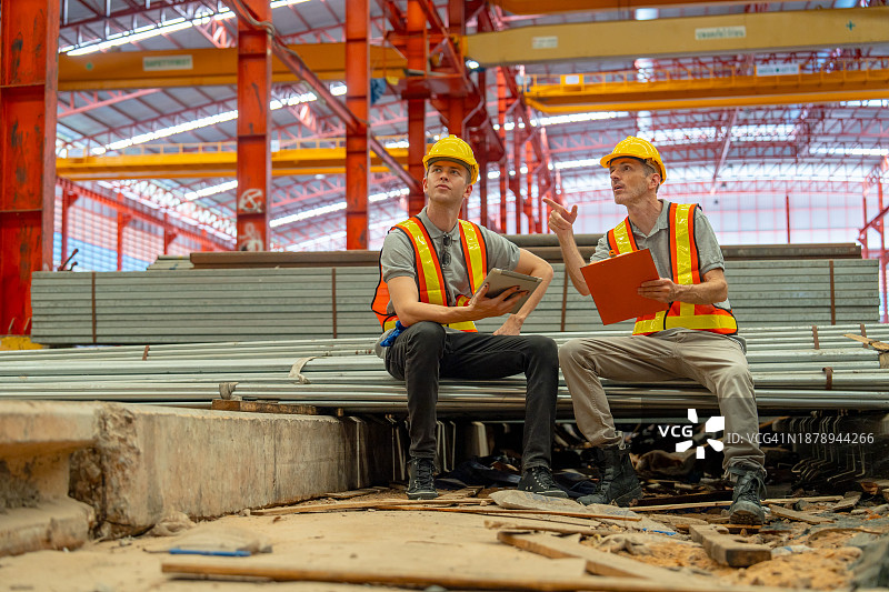 两位工厂工人坐在堆叠的钢管上，在工厂工作区使用平板电脑、纸和文件板讨论工作图片素材