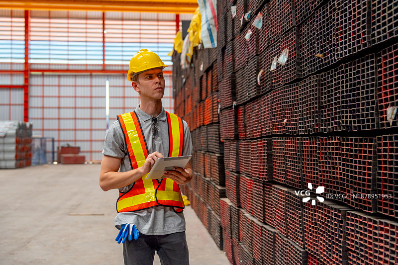 年轻的工厂工人或技术员拿着平板电脑，走向工厂工作区附近的金属管道堆检查产品图片素材