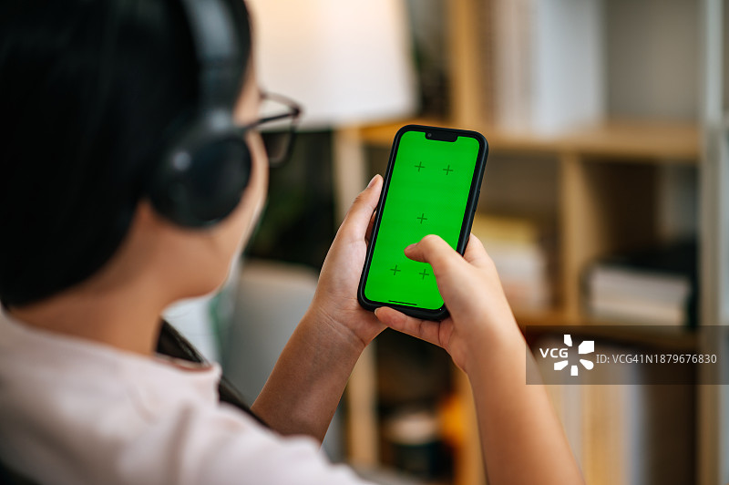 戴着耳机在家中使用带绿屏智能手机的亚洲女商人图片素材