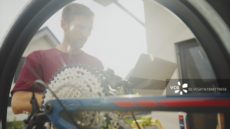 阳光明媚的日子里，男人透过院子里倒置的破损山地自行车看到正在使用笔记本电脑图片素材
