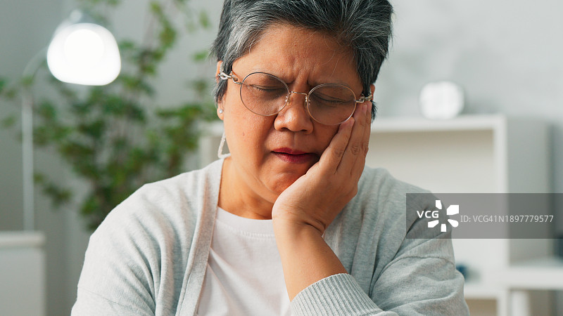 不高兴的亚洲老妇人在家客厅摸着脸颊，有牙齿问题，奶奶患有牙齿敏感，老妇人牙痛图片素材