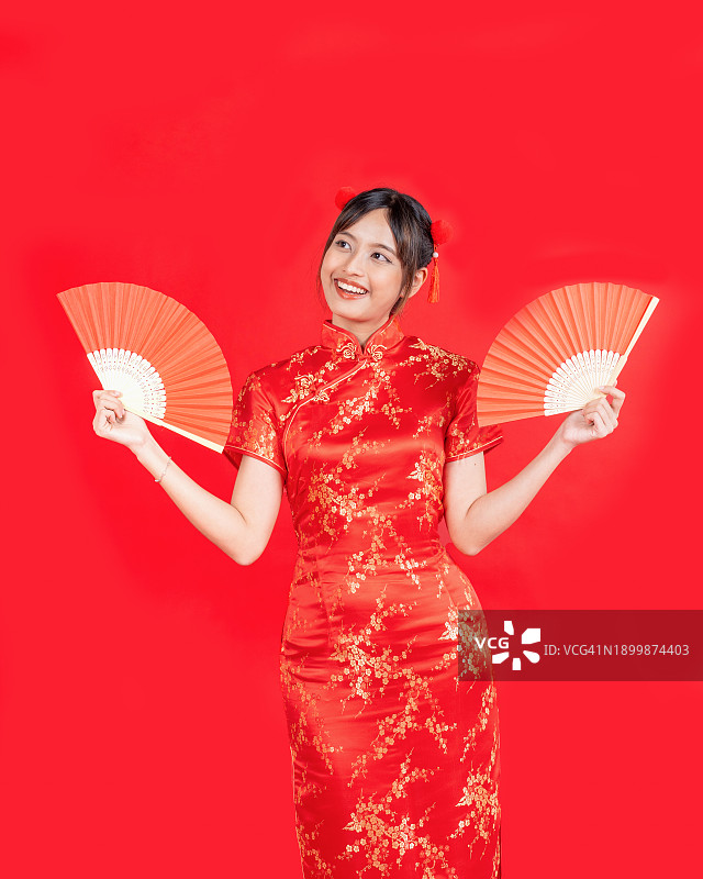 喜庆中国新年，亚洲年轻女性身穿传统旗袍，手持扇子，在红色背景前拍摄图片素材