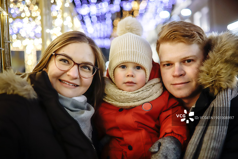 节日街景：年轻夫妇与幼儿在冬季夜晚的闪烁灯光下图片素材