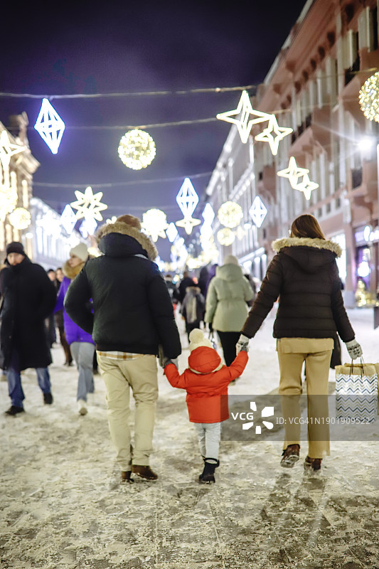 节日街头购物：父母和幼儿一起走在下雪的冬季街道上，手提购物袋，后视图图片素材