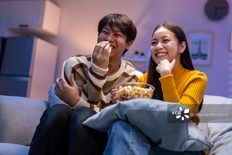 亚洲年轻夫妇在家中沙发上看电视吃爆米花，一起观看喜剧视频，享受快乐约会时光。图片素材