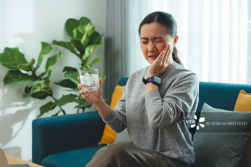 饮用冷水后牙齿敏感，触摸脸颊的亚洲女性，坐在客厅沙发上图片素材