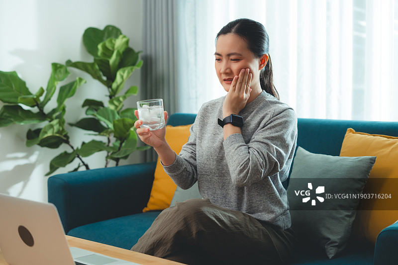 饮用冷水后牙齿敏感，触摸脸颊的亚洲女性，坐在客厅沙发上图片素材