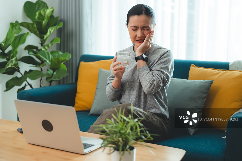 饮用冷水后牙齿敏感，触摸脸颊的亚洲女性，坐在客厅沙发上图片素材