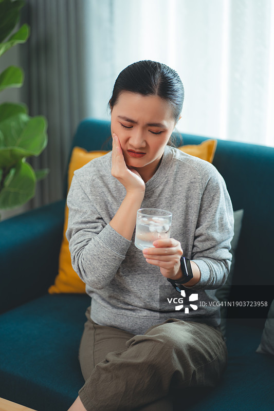 饮用冷水后牙齿敏感，触摸脸颊的亚洲女性，坐在客厅沙发上图片素材