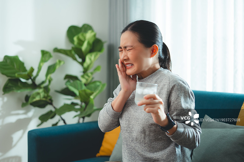 亚洲女性坐在客厅的沙发上，喝了凉水，摸了摸脸颊，就会感到牙齿敏感。图片素材