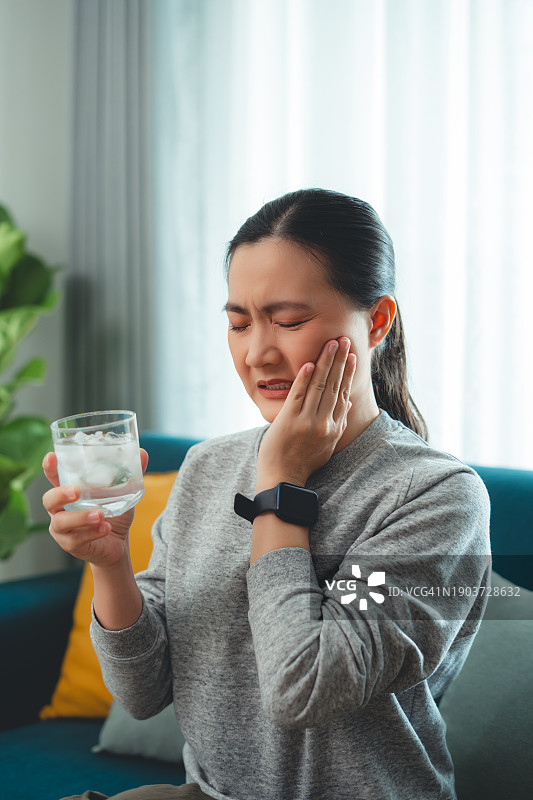 饮用冷水后牙齿敏感，触摸脸颊的亚洲女性，坐在客厅沙发上图片素材