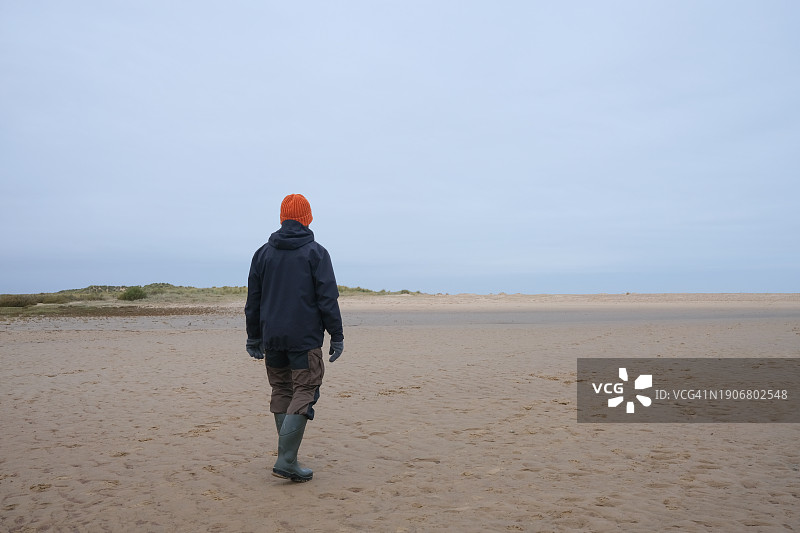 戴帽子和穿雨靴的男子冬天走在荒凉的海滩上图片素材