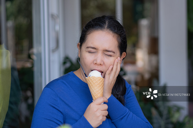 在户外咖啡馆吃冰淇淋时，捂着脸颊缓解牙痛的亚洲女人图片素材