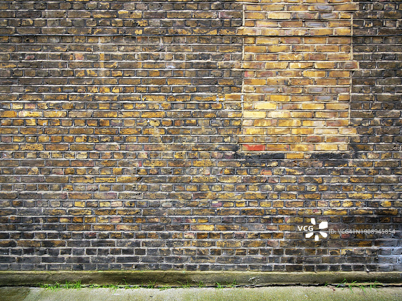 英国伦敦的风化棕黄色砖墙，水泥人行道和杂草图片素材