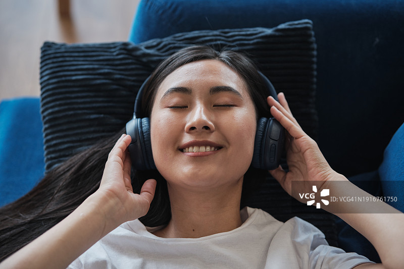 戴着耳机听音乐的快乐放松的亚洲年轻女人在家休息图片素材