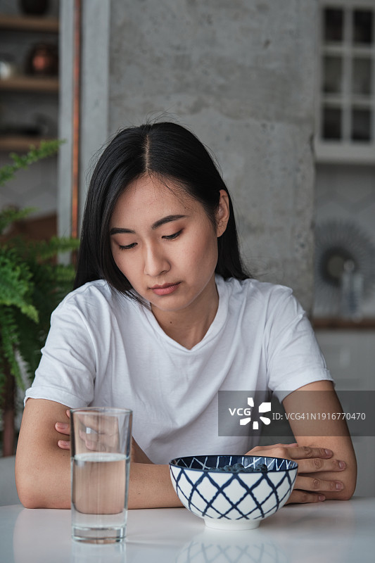 沮丧的亚洲年轻女性坐在厨房餐桌旁，食欲不振，患有贪食症、抑郁症和饮食失调图片素材