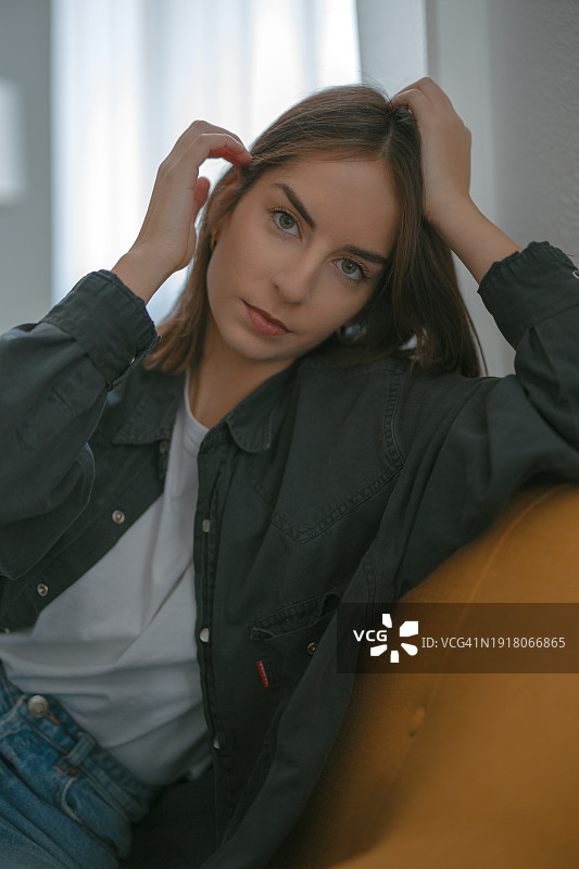 美丽的年轻女子坐在沙发上，看着镜头并将头发别在耳后的肖像图片素材