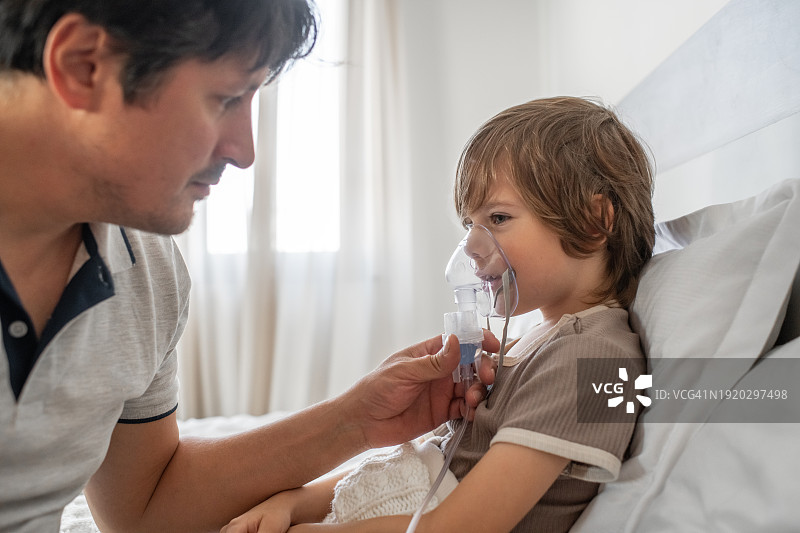 生病儿童在家中床上使用雾化器与父亲一起吸入药物，治疗哮喘、流感、感冒、新冠、支气管炎或肺炎、病毒或感染图片素材