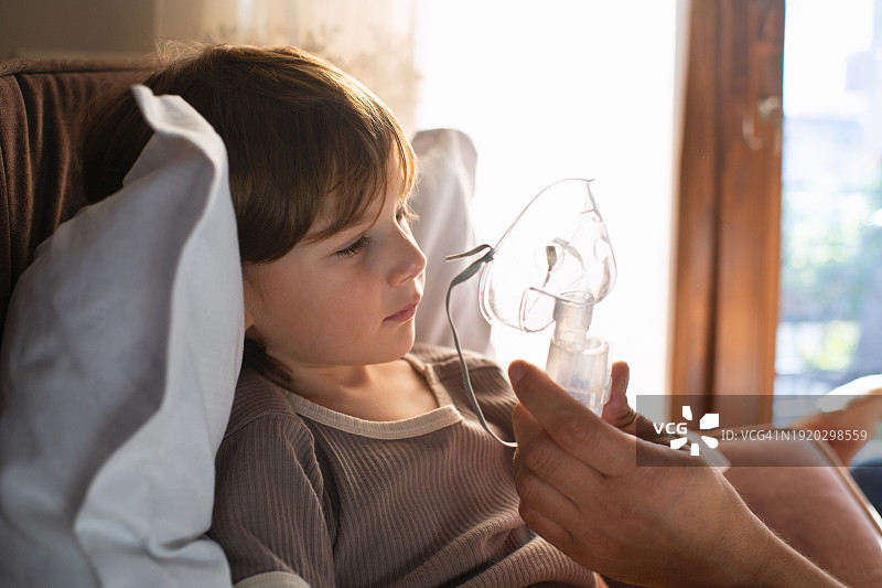 生病儿童在家中床上使用雾化器与父亲一起吸入药物，治疗哮喘、流感、感冒、新冠、支气管炎或肺炎、病毒或感染图片素材