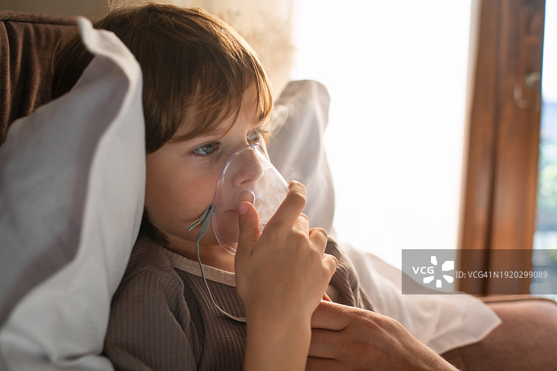 生病儿童在家中床上使用雾化器与父亲一起吸入药物，治疗哮喘、流感、感冒、新冠、支气管炎或肺炎、病毒或感染图片素材