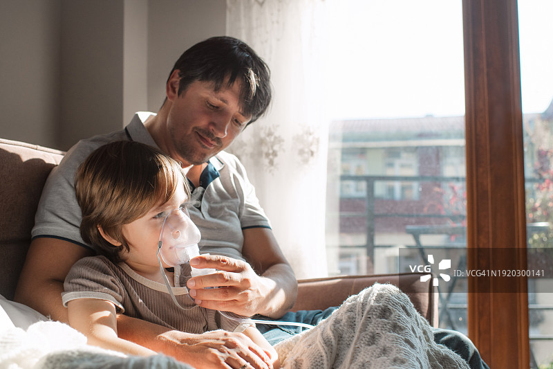 生病的孩子在父亲的帮助下在家用雾化器吸入药物，治疗流感、感冒、新冠、支气管炎或肺炎图片素材