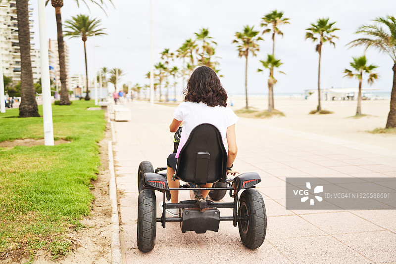 年轻女子在海滩背景的人行道上驾驶电动儿童车背影图片素材