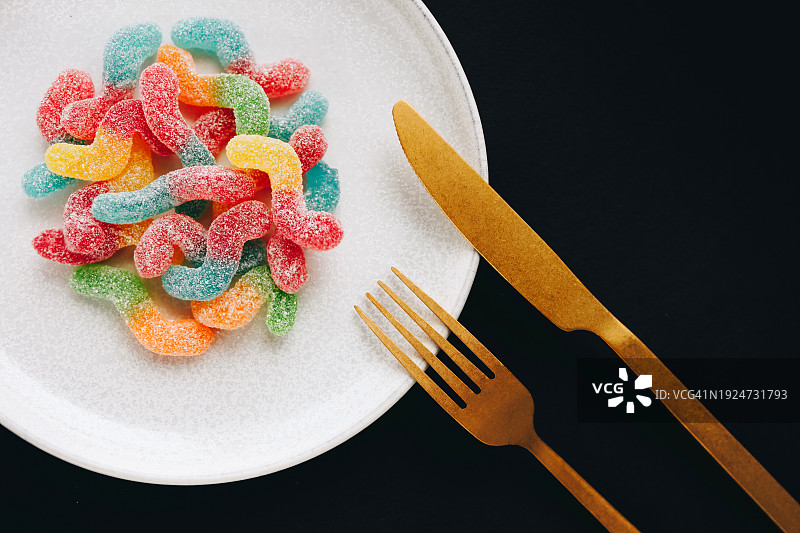 盘子里的彩色水果软糖，配有金色刀叉，黑色背景图片素材