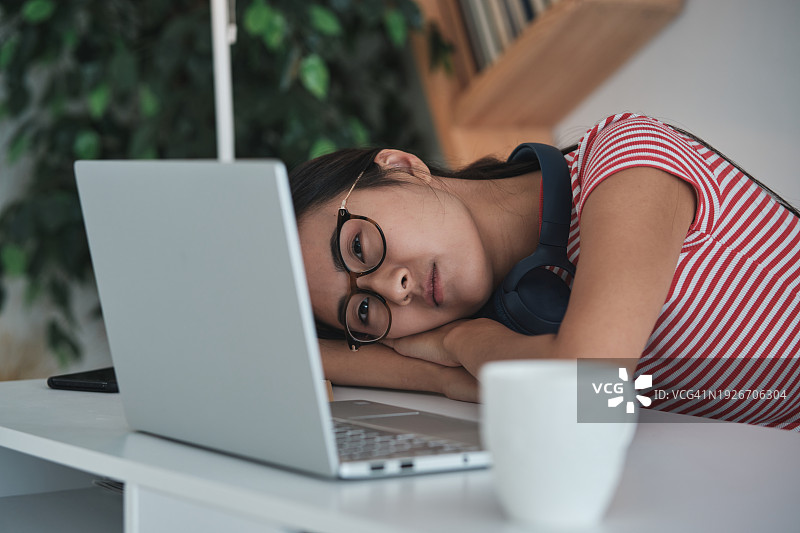 自由职业者倦怠，疲惫不堪的亚洲女性远程工作者躺在桌子上看着笔记本电脑，感到不知所措图片素材