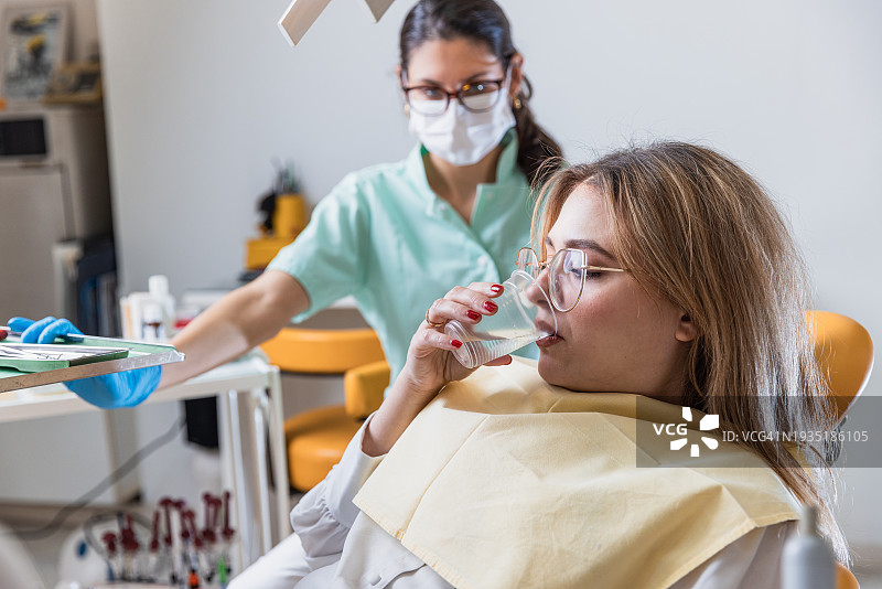 年轻女患者在牙科医生等待继续治疗时，在牙科痰盂上漱口 - 股票照片图片素材