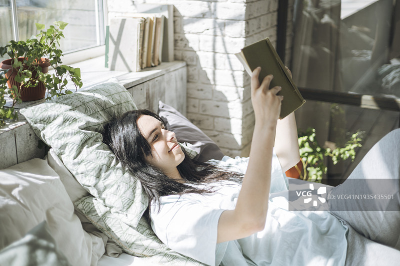 年轻的棕发女子在床上看书。阳光明媚的早晨，开启美好的一天图片素材