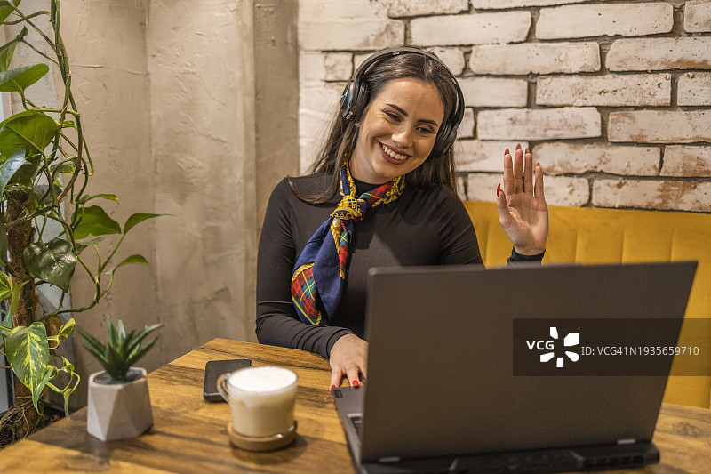 快乐美丽的时尚黑发女人在自助餐厅通过视频通话看着她的笔记本电脑屏幕打招呼图片素材