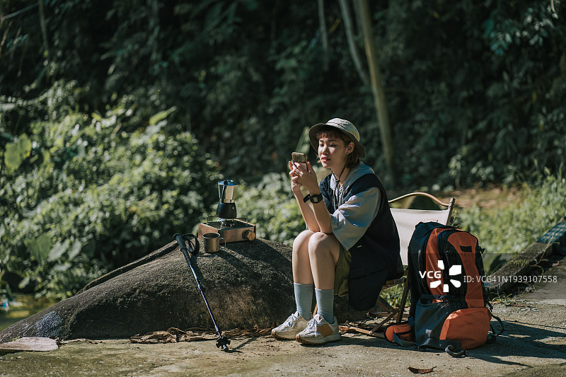 中国亚洲年轻女子在周末早晨徒步旅行后，享受大自然，用智能手机发短信，并在瀑布旁煮咖啡休息图片素材