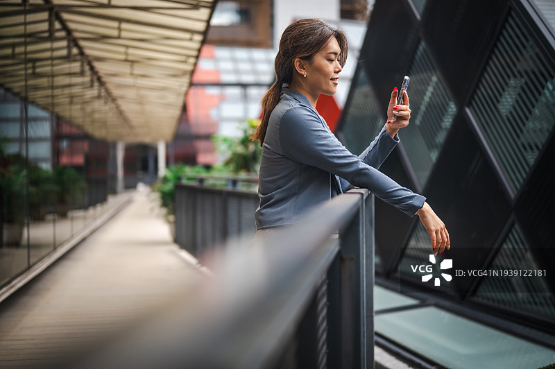 企业风采：一位年轻的亚洲女商人手持智能手机在现代办公室走廊中的迷人肖像图片素材