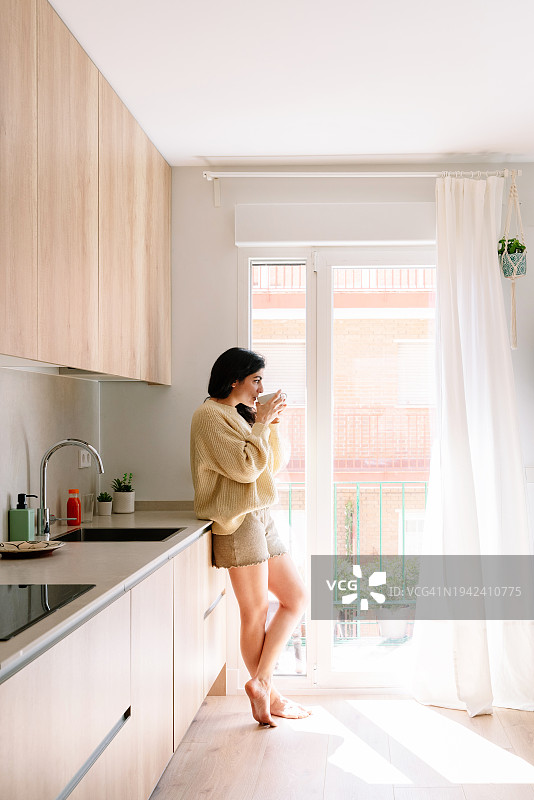 西班牙裔妇女穿着睡衣站在现代木制厨房台面上喝热茶图片素材