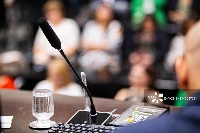 完整的会议室，前景是背对着的人，有麦克风、键盘、电子投票设备、一杯水和一支笔图片素材