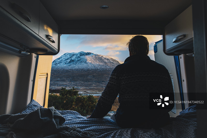 中年男人在他的露营车里的床上欣赏苏格兰高地壮丽的雪山和冬季景色图片素材