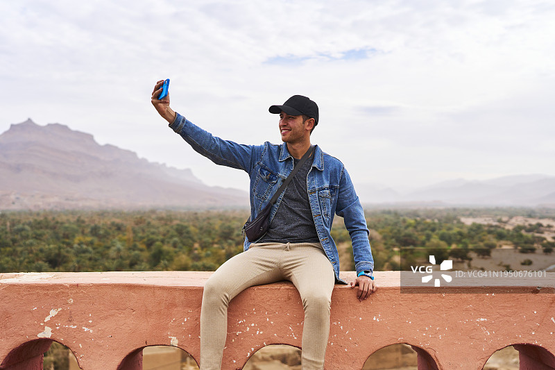 坐在市政厅栏杆上，用手机在马拉喀什沙漠背景下自拍的男游客肖像图片素材