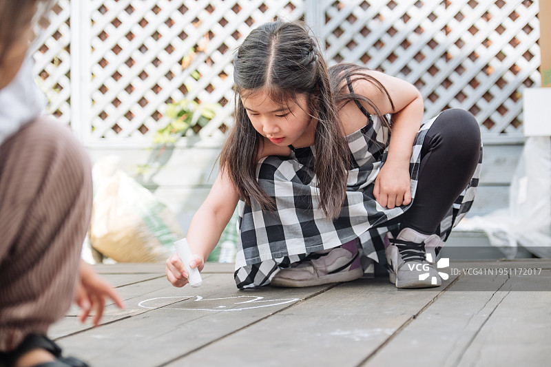 亚洲小女孩和朋友在夏日阳光明媚的日子里用粉笔在户外木地板上绘画图片素材