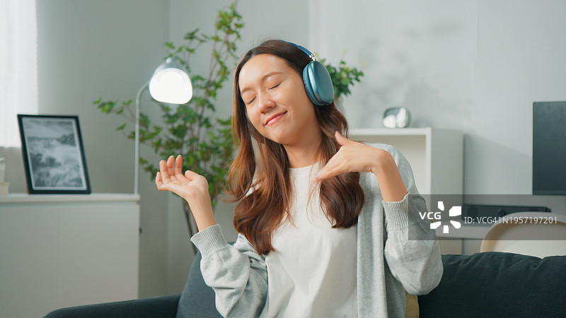 快乐的年轻亚洲女子穿着舒适的衣服，戴着无线耳机在客厅听音乐图片素材