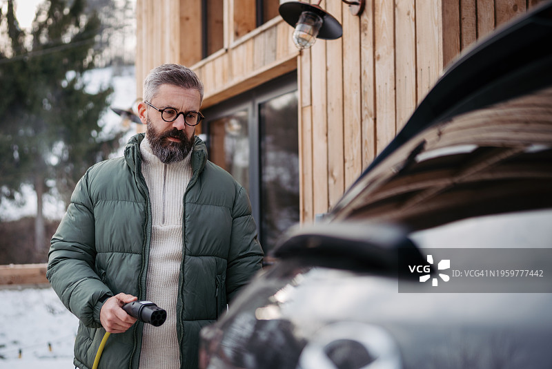 男人在山间小屋前为电动汽车充电，冬季驾驶电动汽车度假图片素材