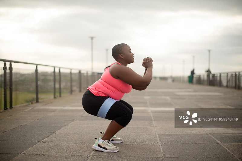 加大码的黑人女性在户外进行深蹲，以达到健康、训练或有氧运动的目的。减肥、运动或在人行道上进行身体锻炼、健康或挑战的非洲女性的轮廓图片素材
