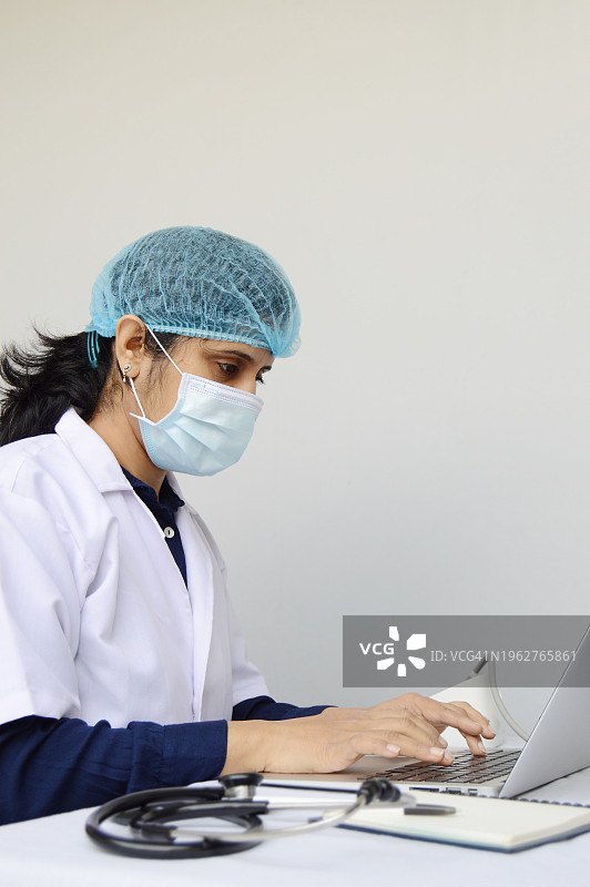 戴着防护口罩、帽子和白色实验服的印度女医生在笔记本电脑上工作，灰色竖版背景，桌上有听诊器，有文字复制空间图片素材