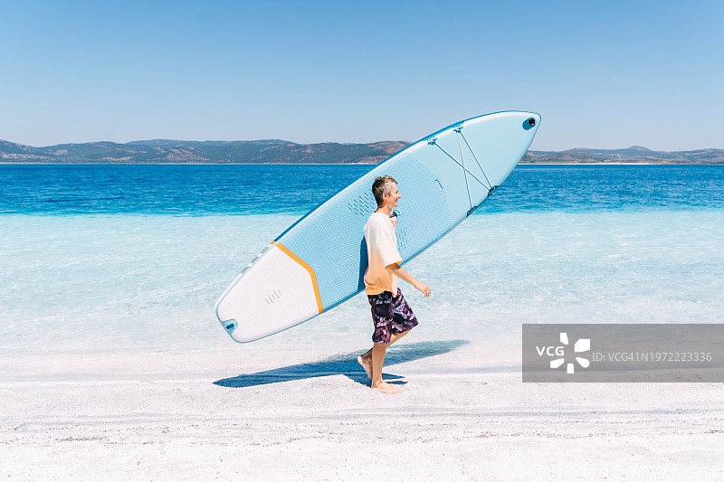 抱着蓝色桨板走在海边的年轻男人图片素材