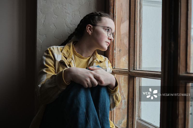 悲伤沮丧的青少年在家中。少女望着窗外，沮丧的孩子坐在窗台上，情绪低落。青少年心理健康图片素材