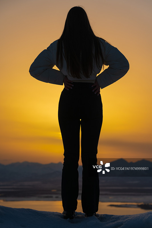 一个无法辨认的少女平静地站在山边，双手叉腰，以美丽的日落天空为背景的剪影后视图图片素材
