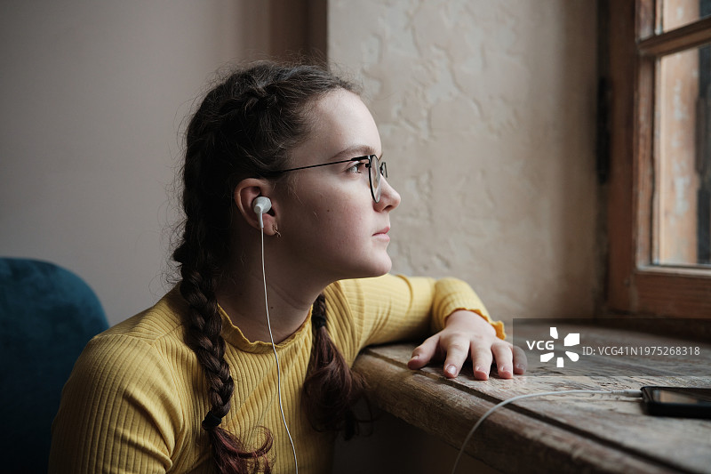 悲伤的少女戴着耳机听音乐，望着窗外，青少年在家感到孤独。青春期的孤独图片素材