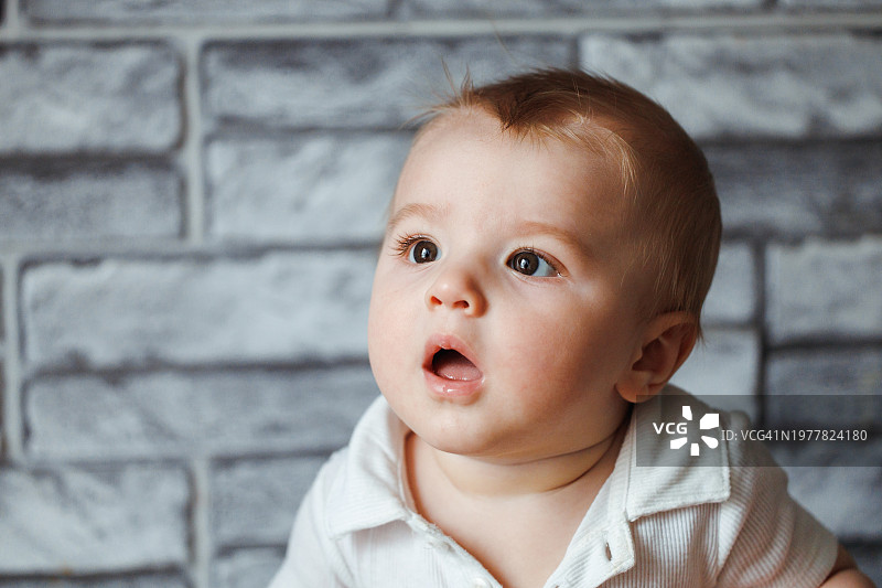 白色衬衫的金发婴儿男孩肖像，背景是模仿砖块的灰色壁纸图片素材